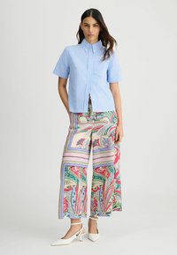 Chemise bleu clair à manches courtes et boutons, avec des rayures verticales ; associée à un pantalon large à motifs paisley colorés et des sandales à talons blanches.