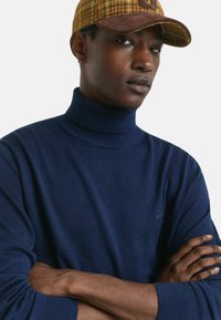 Navyblauwe turtleneck trui van zacht materiaal, met ribbels aan de mouwen. Het model draagt een geel en bruin geruit petje. De achtergrond is eenvoudig wit.