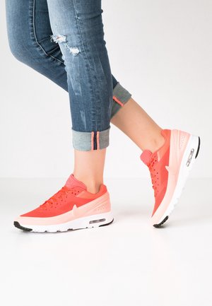 Osoba nosząca podwinięte niebieskie dżinsy i jaskrawo koralowe buty Nike Air Max, stojąca z jedną stopą lekko uniesioną na białej podłodze.