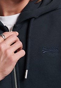 Felpa con cappuccio blu navy con zip frontale, tessuto strutturato e logo ricamato sul petto sinistro. Presenta coulisse e dettagli in metallo.