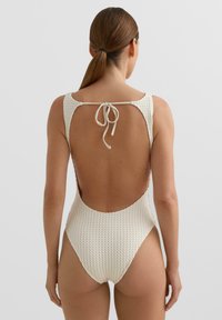 Biely, štruktúrovaný jednodielne plavky s nízkym chrbtom a nastaviteľným zaväzovaním. Má široké ramienka a vysoké vykrojené nožičky.