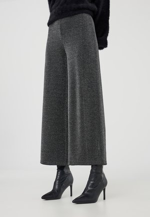 Femme portant un pantalon en maille gris à jambes larges avec des bottines noires à talons aiguilles et pointues, sur fond uni.