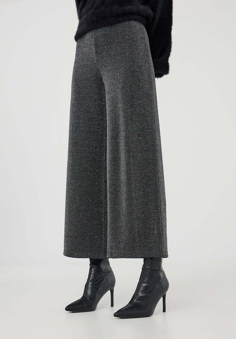 Femme portant un pantalon en maille gris à jambes larges avec des bottines noires à talons aiguilles et pointues, sur fond uni.