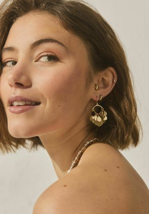 Woman with short brown hair wearing gold disc earrings, a beaded necklace, and a small ear cuff, looking over her shoulder with a slight smile.
