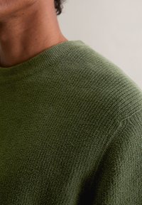 Pulover verde măsliniu cu un design texturat, având un guler rotund și mâneci scurte, evidențiind un aspect moale și confortabil.