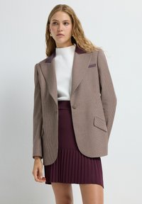 Brauner, gemusterter Blazer mit karminroten Akzenten, einreihiges Design, scharfe Revers und eine Tasche. Kombiniert mit einem plissierten karminroten Rock.