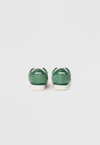 Par čevljev iz zelene semiša je prikazan s hrbtne strani na enobarvni beli podlagi, z belimi podplati in detajli na petah.