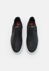 Zapatos tipo slip-on hechos de cuero negro texturizado con paneles laterales elásticos y suelas de goma blanca. El interior presenta una plantilla color burdeos.
