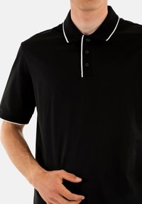 Polo nero con colletto bianco a contrasto e bottoni. Maniche corte, texture liscia, con un piccolo logo nella zona del petto.