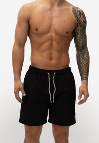 Zwarte zwemboxers met een elastische tailleband en een koord. De stof lijkt licht van gewicht. Opvallende tattoo op de rechterarm.