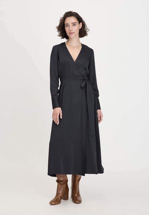 Abito nero avvolgente con maniche lunghe, scollatura a V, cintura in vita, tessuto fluente e una silhouette lunga. Indossato con stivali marroni sopra il ginocchio.