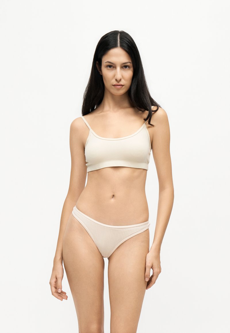 Femme aux longs cheveux foncés, debout face à l’avant, portant un soutien-gorge beige clair et une culotte taille haute assortie sur un fond blanc uni.