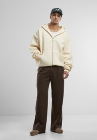 Beige Kapuzenpullover mit Reißverschluss und großer Kapuze, getragen über einem weißen Tanktop, kombiniert mit braunen weit geschnittenen Hosen und braunen Slippern. Dunkle Kappe als Akzent.