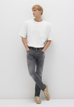 White short-sleeve t-shirt with a pocket, paired with faded grey jeans and beige footwear. Model stands against a plain white background.