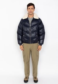 Man staat rechtop met zijn gezicht naar voren, gekleed in een marineblauwe gewatteerde jas met een witte schapenvachtkraag, beige cargobroek en beige wandelschoenen op een witte achtergrond.