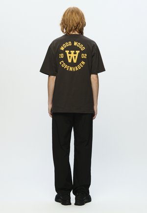 Brown short-sleeve t-shirt featuring yellow graphic text, with "WOOD WOOD COPENHAGEN" and a logo. Black loose-fitting pants and black shoes.