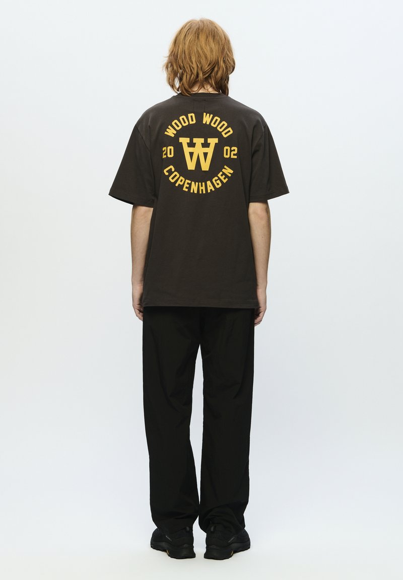 Braunes kurzärmeliges T-Shirt mit gelbem Grafiktext, darauf "WOOD WOOD COPENHAGEN" und einem Logo. Schwarze, locker sitzende Hose und schwarze Schuhe.