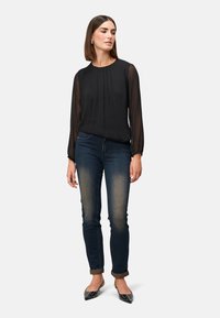 Schwarze gefaltete Bluse mit durchsichtigen Ärmeln, kombiniert mit dunkelblauen Jeans. Das Model trägt schwarze spitze Ballerinas mit Nietenakzenten.