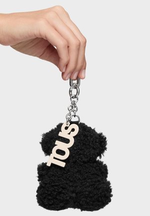 Mano che tiene un portachiavi d'argento con un orsetto nero soffice e una targhetta bianca con la scritta "TOUS" attaccata.
