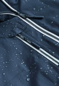 Wodoodporna granatowa kurtka z dwoma suwakami, z białymi szwami, kroplami wody na powierzchni i gładką teksturą.