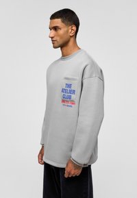 Lichtgrijze sweatshirt met een ontspannen pasvorm, voorzien van een zak en gedurfde blauwe en rode tekst: "THE ATELIER CLUB" zeefdruk op de voorkant.