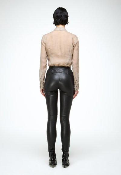 Persoon met kort donker haar, gekleed in een beige shirt met lange mouwen, strakke zwarte leren broek en zwarte enkellaarsjes met hak, staand met de rug naar de kijker.