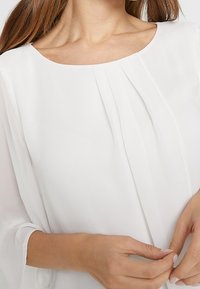 Weißes Blusentop mit rundem Ausschnitt, plissierten Stoffdetails und durchsichtigen langen Ärmeln. Glatte Textur und dezente Drapierung, entworfen für einen lässigen Sitz.