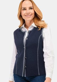 Vrouw in een marineblauwe vest met knopen en grijze afwerking, over een wit shirt, glimlachend, met halflang blond haar.