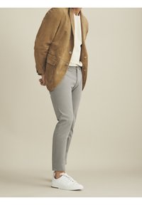 Mies, joka on pukeutunut vaaleanruskeaan mokkajakkuun, valkoiseen neuleeseen, harmaisiin housuihin ja valkoisiin tennareihin, seisoo yksinkertaista beigeä taustaa vasten.