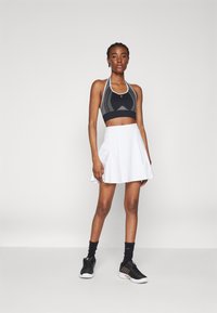 Svart atletisk crop-top med halterneck-design, kombinert med et hvitt, utsvingt skjørt. Svarte joggesko med teksturerte detaljer og svarte sokker.