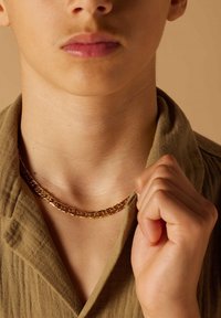 Gouden kettinghalsketting die op de nek rust, met schakels met een gladde afwerking. De kleding van het model is gemaakt van een textuurrijke olijfgroene stof.