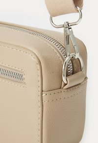 Sac en cuir beige avec surface texturée, zip argenté et quincaillerie en métal. Dispose d'une sangle amovible et d'une petite poche avant.