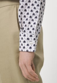 Witte shirt met marineblaue palmbomenpatroon, met een gesloten manchet en zachte stof; gecombineerd met beige broeken, wat een casual stijl uitstraalt.
