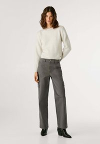 Vrouw met golvend bruin haar, gekleed in een witte trui met lange mouwen, grijze cargobroek en zwarte haklaarzen, staande tegen een effen achtergrond.