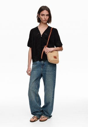 Mujer con blusa negra bordada, vaqueros azules holgados, sandalias, sosteniendo un pequeño bolso bandolera tejido con correa de cuero marrón.