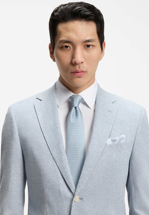 Uomo con abito azzurro chiaro a trama, camicia bianca e cravatta blu a motivi, di fronte a sfondo neutro.