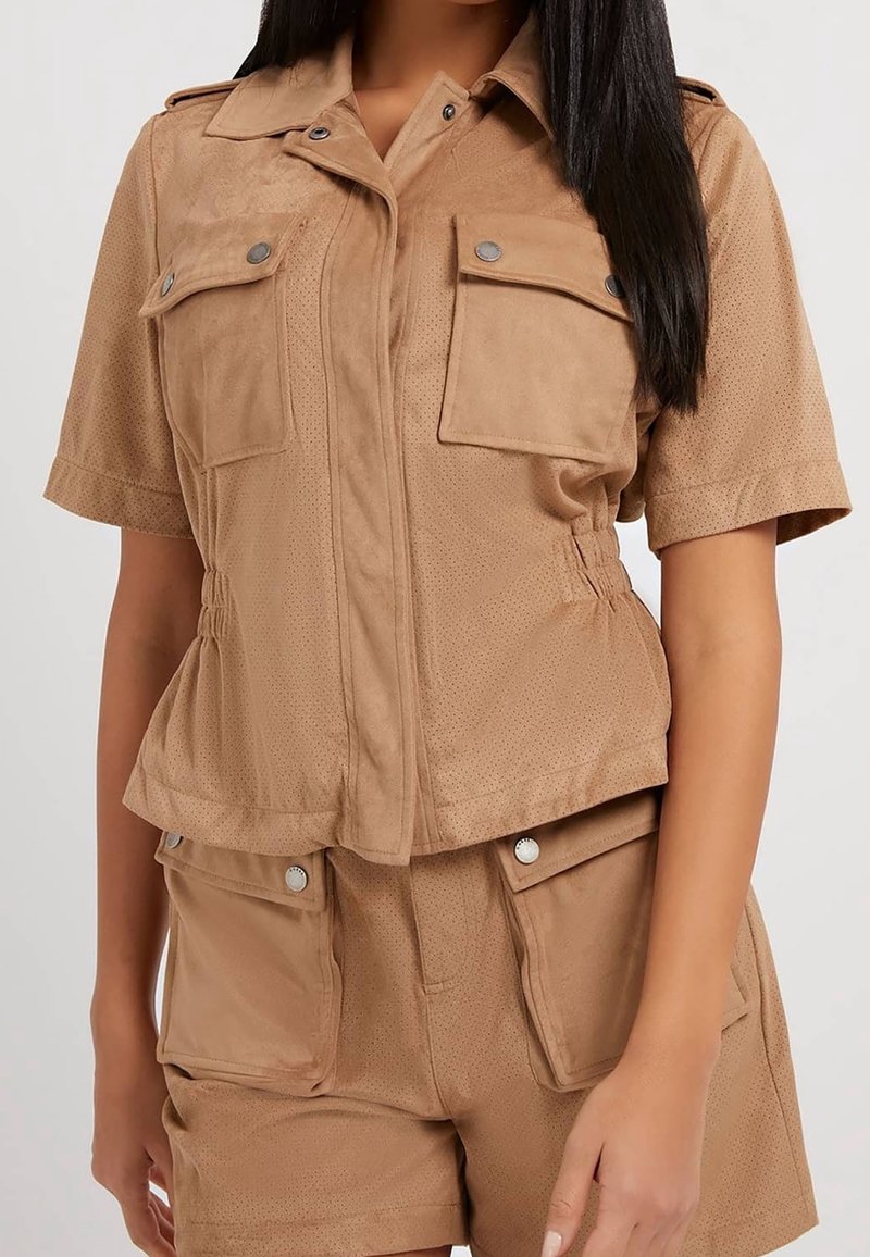 Femme portant une veste beige à manches courtes avec quatre poches à rabat et un short assorti sur un fond uni.