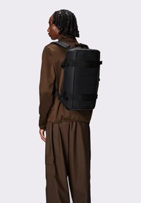 Zaino nero dalla forma rettangolare, con spallacci regolabili e diversi cinturini esterni. Indossato da una persona con una maglia a maniche lunghe marrone e pantaloni.