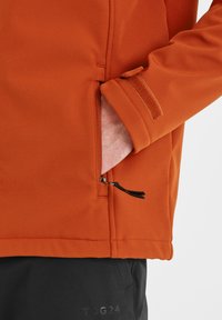 Orange jacket with a soft texture, featuring a side zip pocket and adjustable Velcro cuff. Worn over black pants with logo.