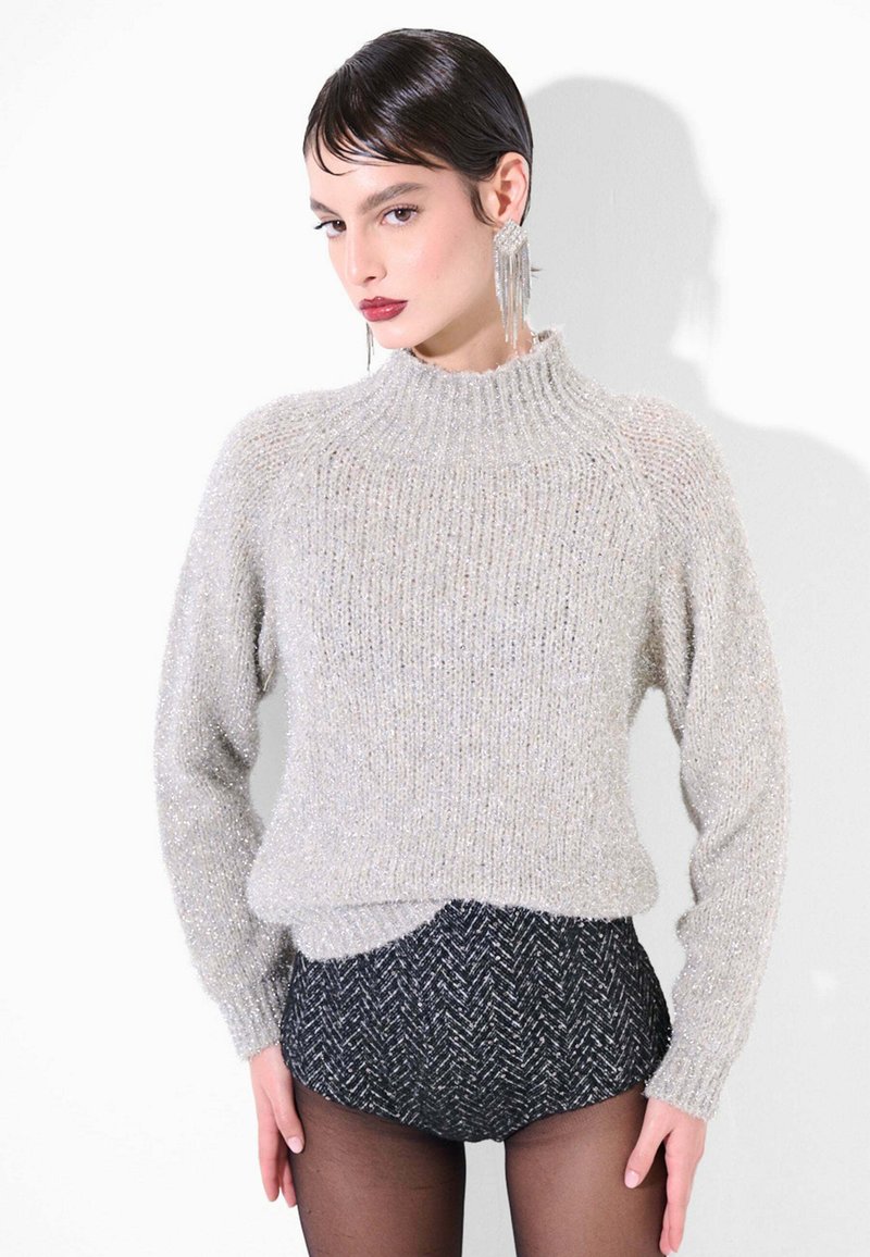 Maglione grigio chiaro a coste con collo alto e texture morbida e scintillante. Abbinato a pantaloncini neri con motivo a spina di pesce. La modella indossa lunghe orecchini pendenti.