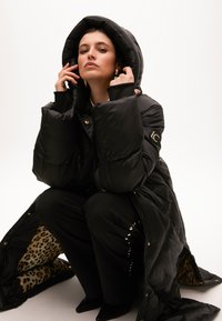 Černá prošívaná bunda s velkou kapucí, s podšitím v leopardím vzoru. Model má na sobě černé skládací kalhoty a černé špi čaté boty.