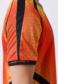 Manche d'un maillot de sport dégradé orange et jaune avec une bande noire et le texte "PROJECT X PARIS" sur le poignet, porté sur le bras d'une personne.
