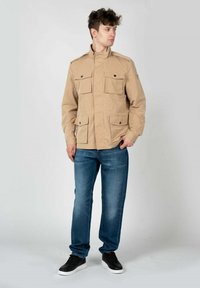 Giacca beige con colletto alto e quattro tasche frontali, abbinata a jeans blu e sneakers nere. Design semplice con accenti minimi.