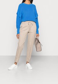 Blå ribbad stickad tröja med kantiga axlar, parat med beiga spetsade byxor med dragsko. Vita sneakers och en beige handväska fulländar looken.