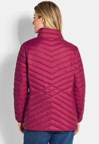 Vrouw met blond haar, gekleed in een magenta gewatteerde jas en blauwe jeans, met haar rug naar de camera gekeerd, tegen een effen lichte achtergrond.