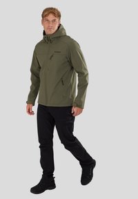 Olivengrøn softshell-jakke med hætte, lynlås-lommer og slim fit, parret med sorte bukser og robust sort fodtøj.