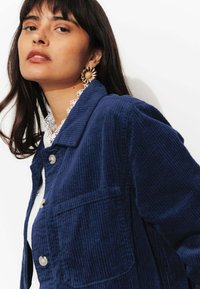 Veste en velours côtelé bleu marine avec un design à col, dotée d'une poche avant et de boutons en argent. Texture chaude ; portée sur un top en dentelle blanc.