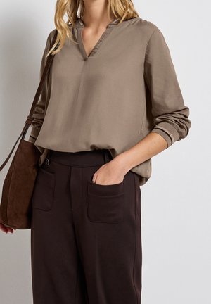 Vrouw draagt een taupekleurige blouse met lange mouwen met manchetdetail, zwarte hoge taillebroek met voorzakken en draagt een bruine schoudertas.