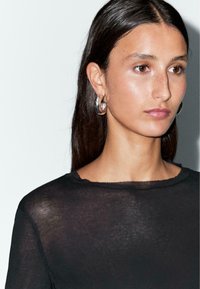 Black sheer long-sleeve top with a round neckline and frayed edges, paired with large silver hoop earrings. Smooth texture.