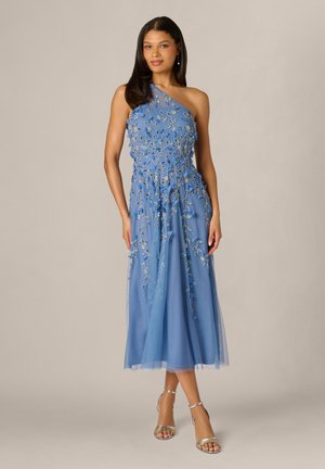 Vrouw draagt een blauwe midi-jurk met een open schouder, bloemrijke borduursels en zilveren sandalen met hoge hak, staand tegen een effen achtergrond.